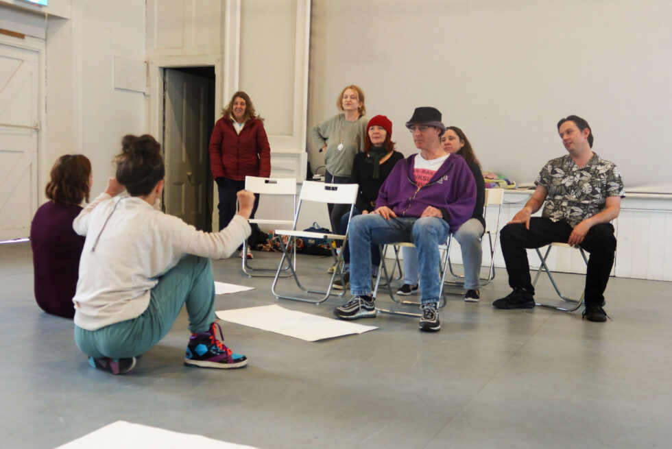 Inside a rehearsal room, eight people sit and stand poised for performance.