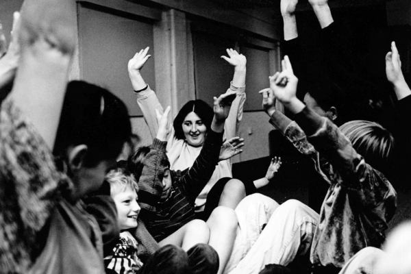 A group of children and young people sit with their hands up in the air, they are smiling and cheerful.