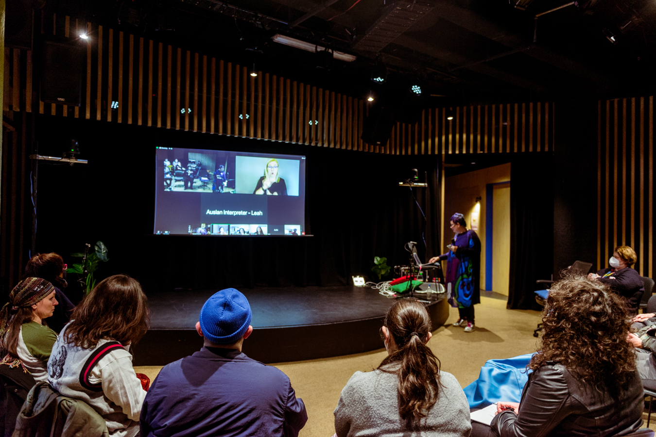 A group sit facing a large projection with squares of people streaming in. A presenter is looking at a computer.