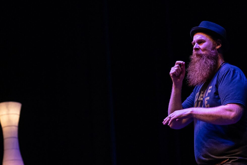 Ramas McRae in performance in a dark theatre space, with two lights to the left of the image.