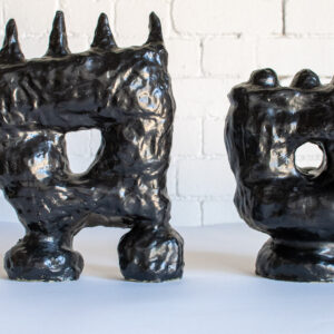 These two sculptures are black. They both sit on two stands with a larger box-like shape sitting on top. There is a hole in the middle that allows you to see through each sculpture. On top are spikes on one and round blobs on the other.