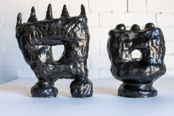 These two sculptures are black. They both sit on two stands with a larger box-like shape sitting on top. There is a hole in the middle that allows you to see through each sculpture. On top are spikes on one and round blobs on the other.