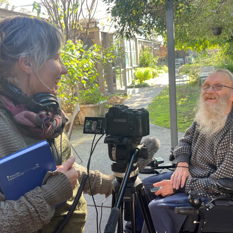 Two people in a backyard. Leonie filming artist Peter Strang, they are both smiling.