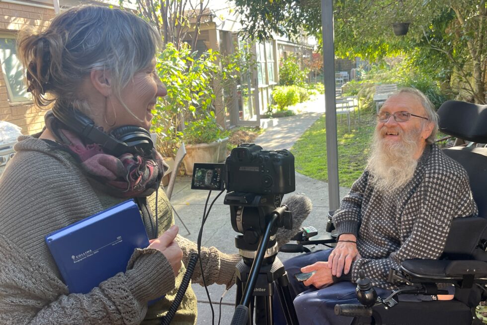 Two people in a backyard. Leonie filming artist Peter Strang, they are both smiling.