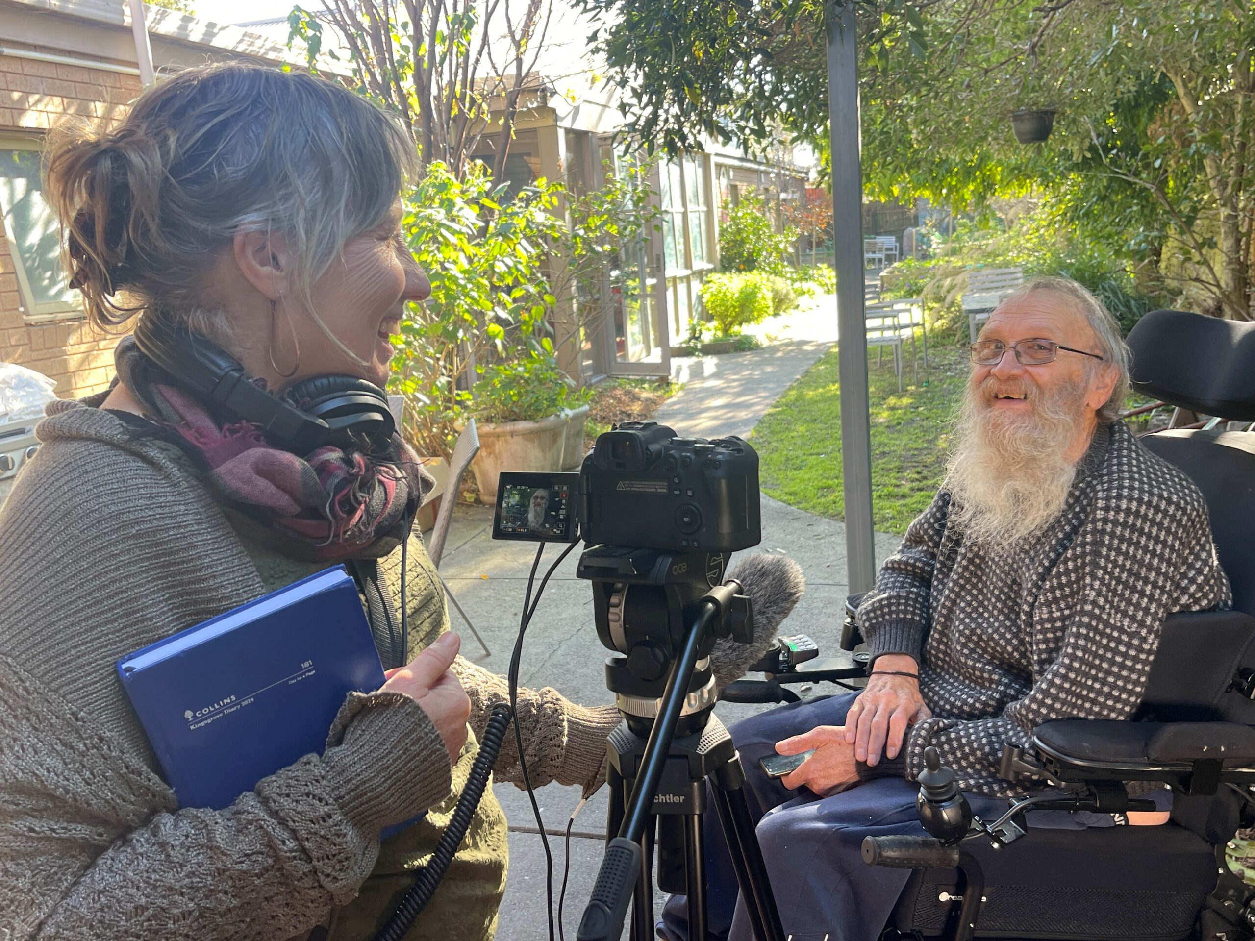 Two people in a backyard. Leonie filming artist Peter Strang, they are both smiling.