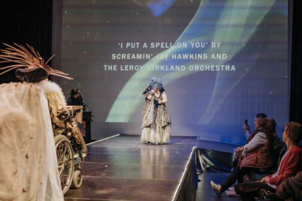 Two people in costumes on a runway stage with audience members seated side of stage. A projection behind them reads 