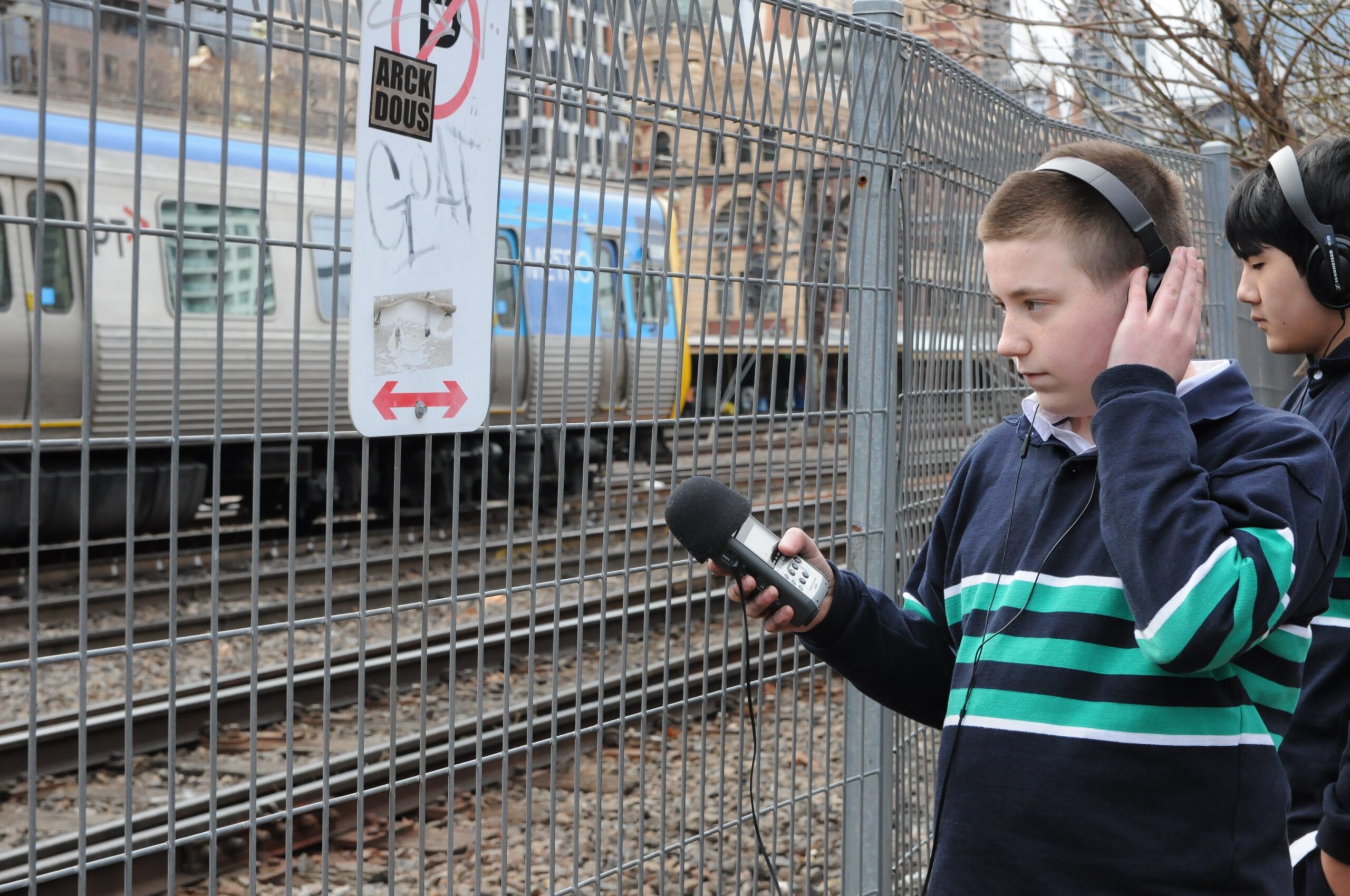 Young person wearing headphones and holding a Zoom recorder towards train tracks.