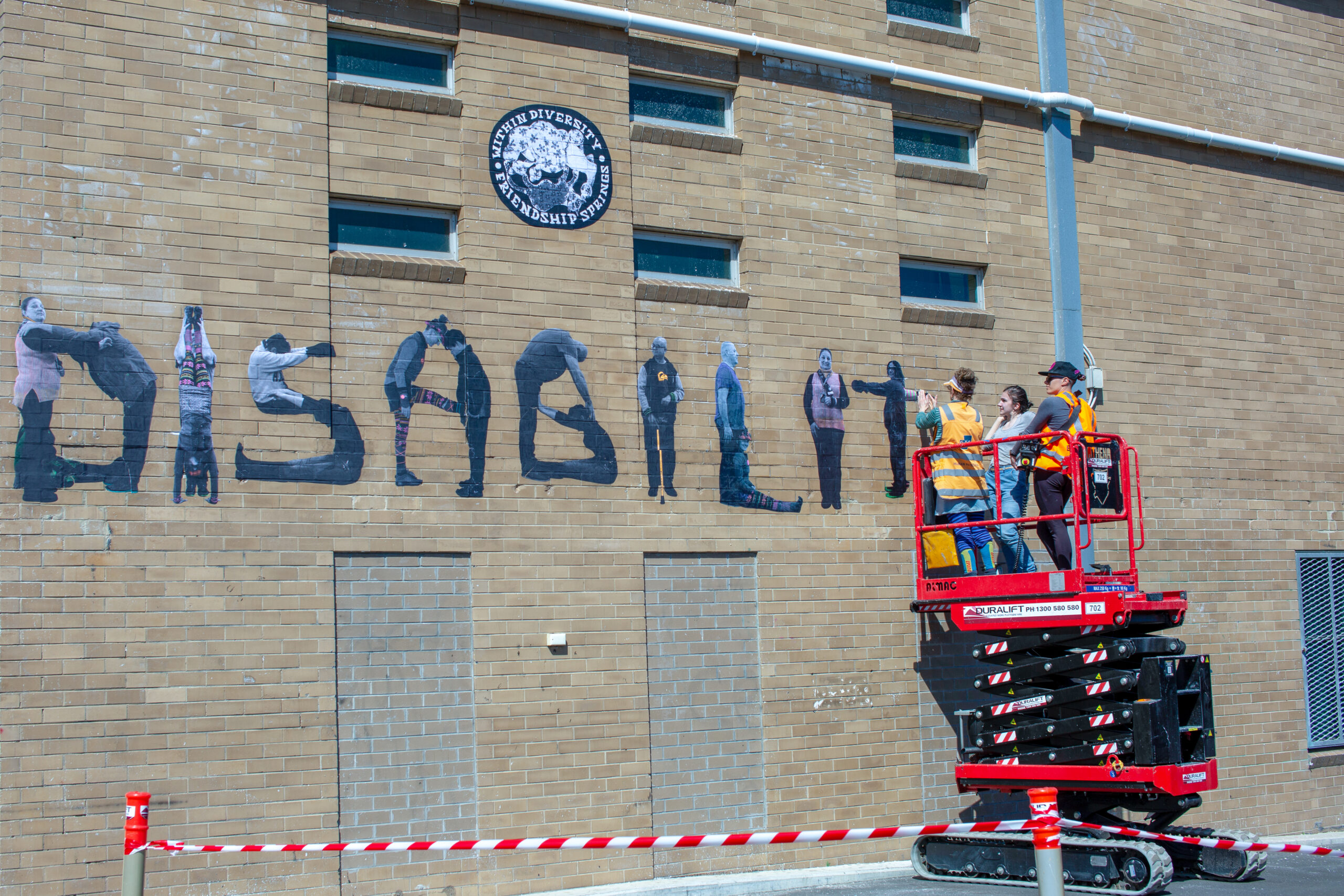 People on a scissor lift installing a mural on a brick building with the word 