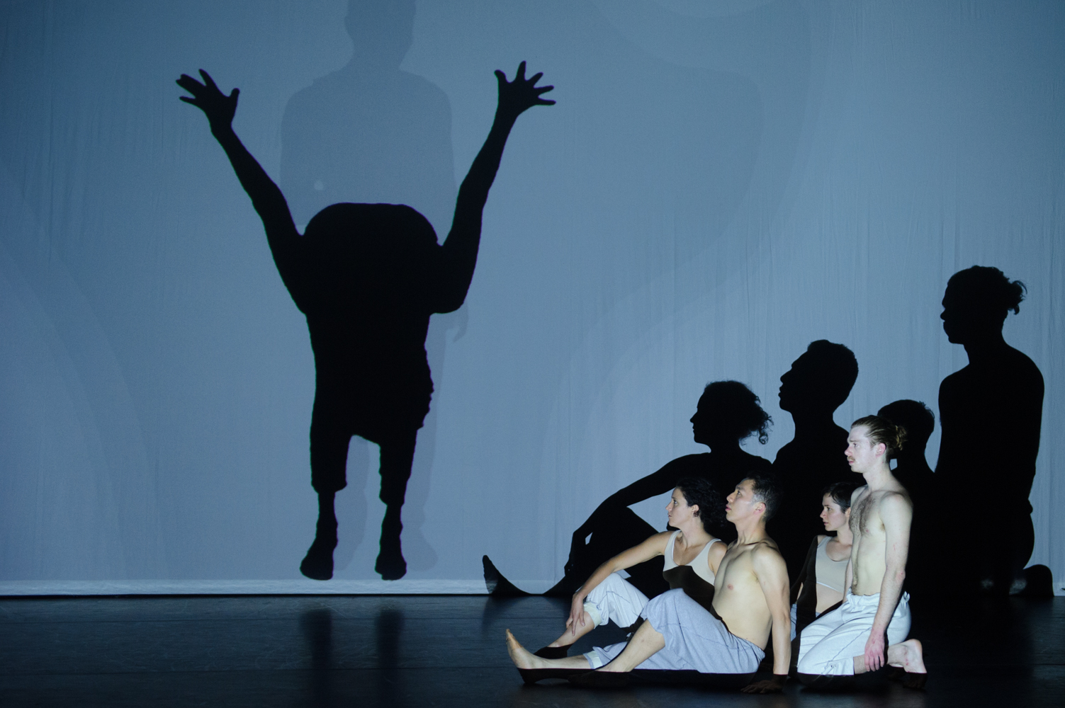 Performers seated and kneeling on stage, their shadows projected behind them. Another performer's shadow creates a unique shape as the others look on.