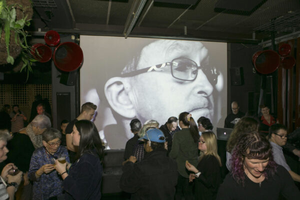 A crowd of people mingling in a room with a large projector screen. A close up of a person's face.